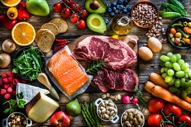 Food backgrounds: top view of a rustic wooden table filled with different types of food. At the center of the frame is a cutting board with beef steak and a salmon fillet and all around it is a large variety of food like fruits, vegetables, cheese, bread, eggs, legumes, olive oil and nuts. DSRL studio photo taken with Canon EOS 5D Mk II and Canon EF 70-200mm f/2.8L IS II USM Telephoto Zoom Lens